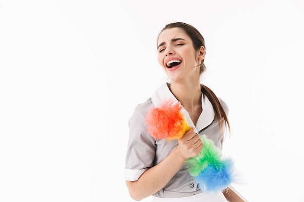 Foto de empregada doméstica feliz dos anos 20, vestida de uniforme, segurando um espanador colorido, enquanto fazia trabalhos domésticos e limpava a sala, isolada sobre uma parede branca