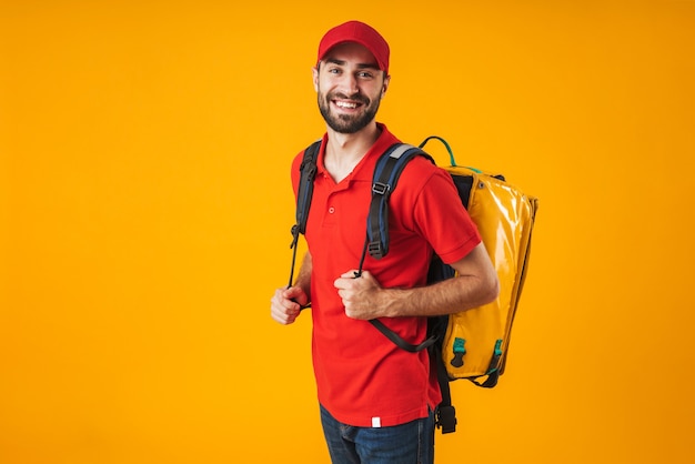 Foto de entregador sorridente com uniforme vermelho carregando uma mochila com comida para viagem isolada sobre o amarelo