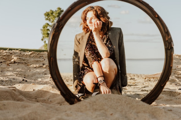 Foto de estilo de vida atmosférica ao ar livre da bela jovem de cabelos escuros no vestido e jaqueta posando na praia no reflexo do espelho.