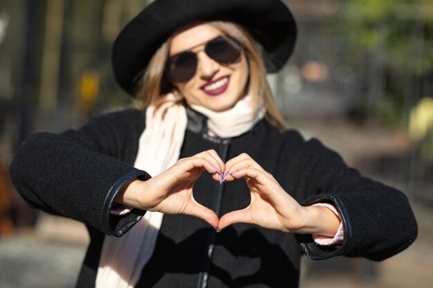 Foto de estilo de vida: modelo loira descolada usando chapéu, mostrando o símbolo do coração na rua