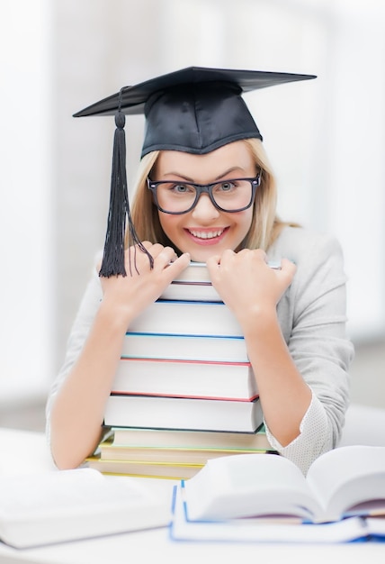 Foto de estudante feliz no chapéu da formatura com uma pilha de livros