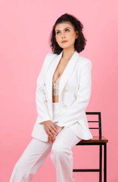 Foto de estúdio closeup retrato de empresária bem sucedida de penteado encaracolado sexy asiático em terno casual elegante branco com sutiã top de lingerie de renda sentado na cadeira alta posando em fundo rosa
