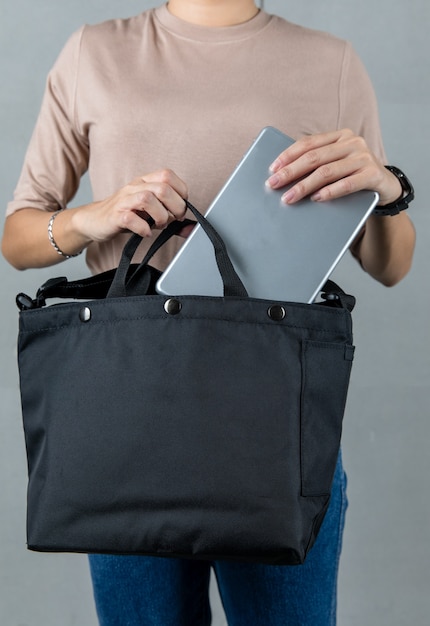 Foto de estúdio de closeup de modelo feminino em camisa de manga comprida marrom, colocando o laptop de prata no bolso do compartimento da bolsa unissex urbana da moda em fundo cinza.