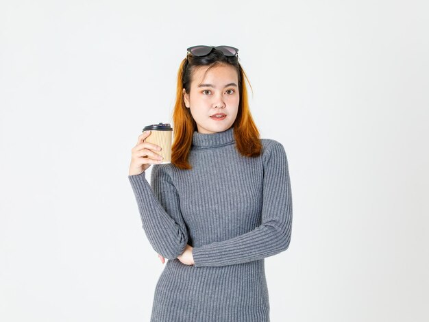 Foto de estúdio de closeup retrato de modelo feminino jovem asiático em vestido de gola alta cinza usar lentes pretas na cabeça em pé sorrindo olhar para a câmera segurando a xícara de café quente braço cruzado sobre fundo branco.