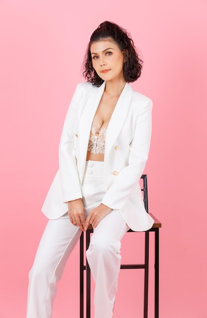 Foto de estúdio de corpo inteiro de retrato de empresária bem-sucedida de penteado encaracolado sexy asiático em terno casual elegante branco com sutiã top de lingerie de renda sentado na cadeira alta posando em fundo rosa