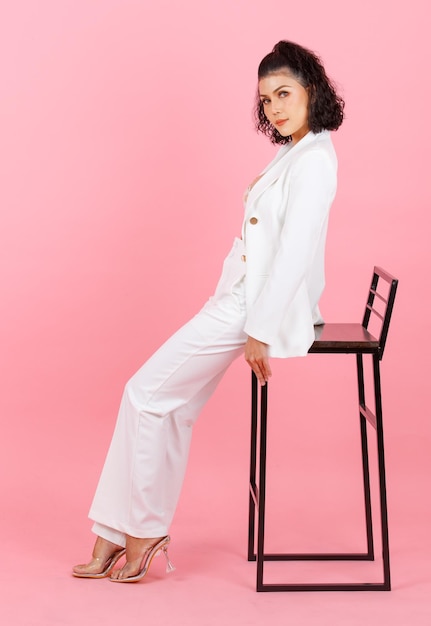 Foto de estúdio de corpo inteiro de retrato de empresária bem-sucedida de penteado encaracolado sexy asiático em terno casual elegante branco com sutiã top de lingerie de renda sentado na cadeira alta posando em fundo rosa