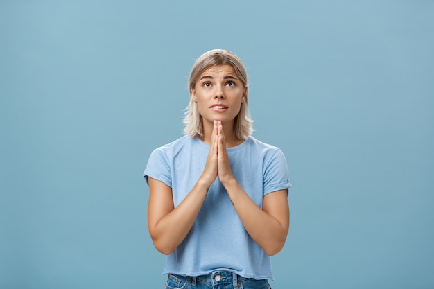 Foto de estúdio de esperançosa garota sonhadora com rosto atraente e cabelo loiro de mãos dadas para rezar perto do corpo, olhando esperançosamente com fé, orando ou fazendo um desejo sobre a parede azul