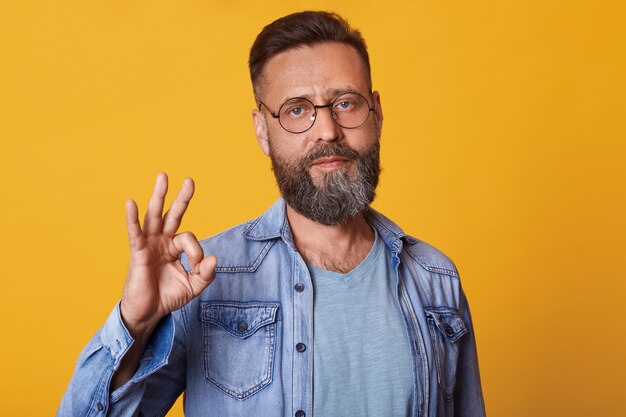 Foto de estúdio de homem bonito com barba, vestindo jaqueta jeans, camiseta cinza e óculos, tem expressão facial positiva