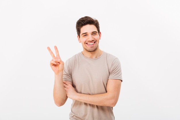 Foto de estúdio de homem bonito, mostrando o símbolo da paz na câmera com um sorriso sincero, isolado sobre a parede branca