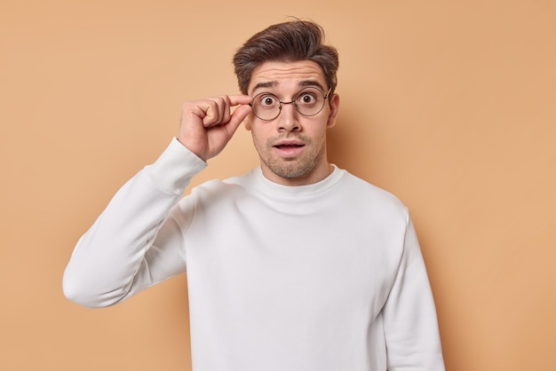 Foto de estúdio de homem moreno espantado mantém a mão na borda dos óculos olha para algo inacreditável, não posso acreditar que seus olhos usam um macacão branco casual isolado sobre um fundo bege. conceito omg