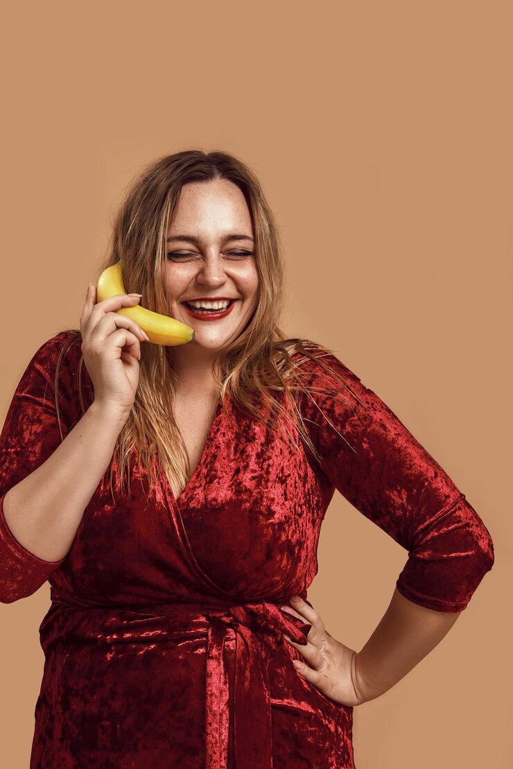 Foto foto de estúdio de humor lúdico de uma mulher sorridente em tamanho grande em um vestido de veludo vermelho segurando uma banana