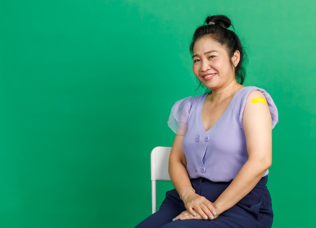Foto de estúdio de mulher asiática de meia idade sentar sorriso e mostrar atadura de gesso amarelo no braço depois de receber a vacinação de coronavírus covid 19 pelo médico na clínica sobre fundo verde.