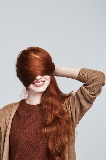 Foto de estúdio de mulher ruiva alegre e jovem brincando com seus cabelos e sorrindo em pé contra um fundo cinza. Conceito de beleza. Felicidade