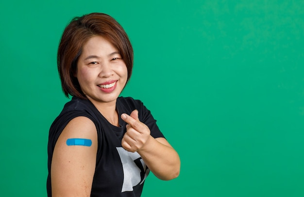 Foto de estúdio de paciente feminino de meia idade asiático sentar sorriso olhar para câmera mostrar mini coração sinal de mão e bandagem de gesso azul no braço após receber a vacinação de coronavírus covid 19 sobre fundo verde.