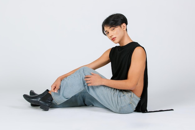 Foto de estúdio de recorte isolado de retrato de asiático jovem sexy luxo glamour magro elegante LGBTQ modelo masculino gay em preto sem manga camisa jeans botas de couro sentado no chão posando em fundo branco