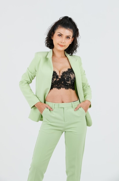 Foto de estúdio de recorte isolado de retrato de empresária de moda de cabelo encaracolado sexy asiática de terno verde com sutiã top de lingerie de renda preta em pé posando colocar as mãos nos bolsos das calças no fundo branco
