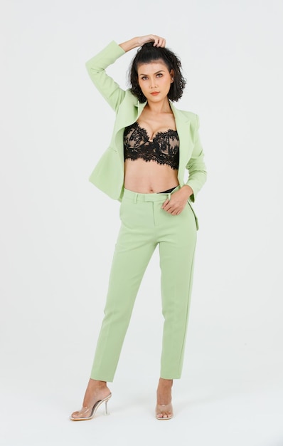 Foto de estúdio de recorte isolado de retrato de empresária de moda de cabelo encaracolado sexy asiática de terno verde com sutiã top de lingerie de renda preta em pé posando colocar as mãos nos bolsos das calças no fundo branco