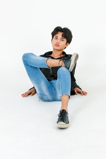 Foto de estúdio de recorte isolado retrato do jovem asiático LGBTQ glamour gay modelo masculino bonito em jeans de camisa preta casual e tênis sentado pernas cruzadas posando gesticulando no chão no fundo branco