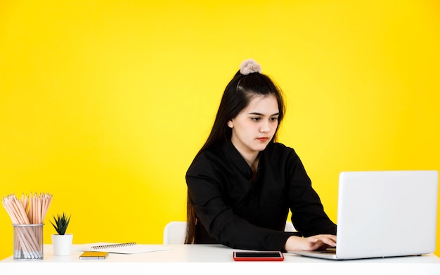 Foto de estúdio de retrato de asiático mal-humorado estressado deprimido empresária empresária em roupa preta, sentado trabalhando horas extras com laptop na mesa do local de trabalho em fundo amarelo.