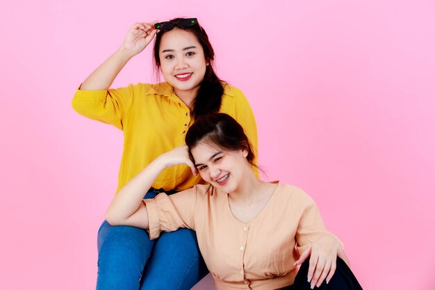 Foto de estúdio de retrato de dois asiáticos jovem alegre lindo rabo de cavalo feminino amigas ou irmãs modelo em roupa casual com aparelho, óculos de sol e chapéu sorrindo olhar para a câmera posando em fundo rosa.