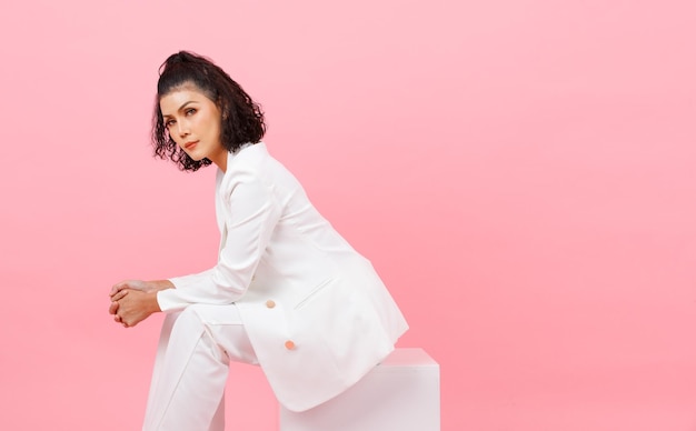 Foto de estúdio de retrato de jovem asiático penteado encaracolado sexy empresária de sucesso profissional em moda branca terno casual sentado posando de mãos dadas juntos na caixa quadrada no fundo rosa