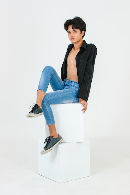 Foto de estúdio de retrato do jovem asiático LGBT gay bissexual homossexual em topless modelo masculino elegante em camisa preta casual e jeans sentado em caixas quadradas posando olhar para a câmera no fundo branco