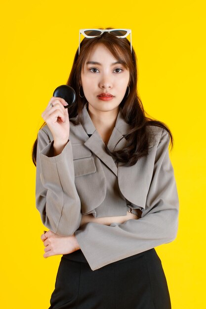 Foto de estúdio de retrato do modelo de adolescente feminino de moda urbana jovem asiático usando maquiagem casual top crop top camisa em pé braço cruzado segurando o estojo de pó olhar para a câmera no fundo amarelo.