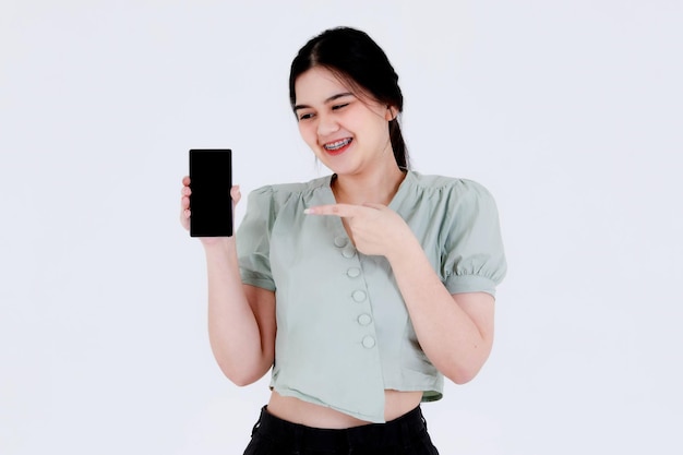 Foto de estúdio de retrato do modelo feminino jovem adolescente asiático com penteado de rabo de cavalo, usando aparelho dentário e equipamento superior de corte apontando tela em branco smartphone em fundo branco.