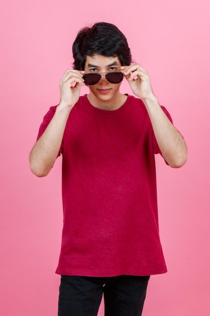 Foto de estúdio de retrato do modelo masculino de moda jovem adolescente urbano asiático em camiseta casual de roupa de estilo de rua e jeans preto com óculos de sol aviador em pé posando olhar para a câmera no fundo rosa