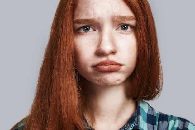 Foto de estúdio de ruiva com camisa quadriculada, fazendo careta em pé contra um fundo cinza. emoções humanas