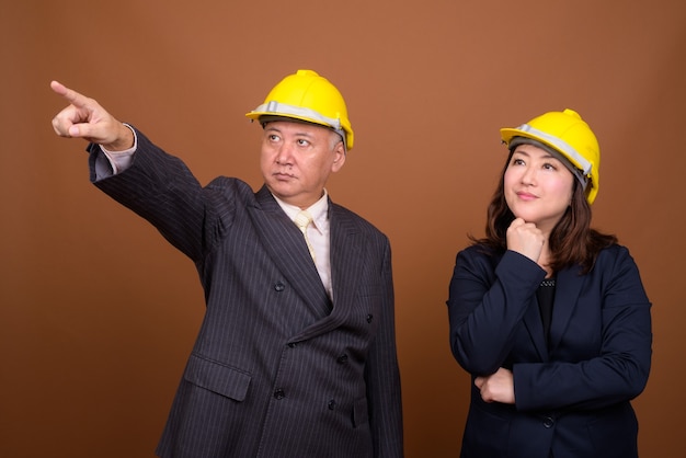 Foto de estúdio de um empresário japonês maduro e uma mulher de negócios japonesa madura usando capacete de segurança juntos contra um fundo marrom