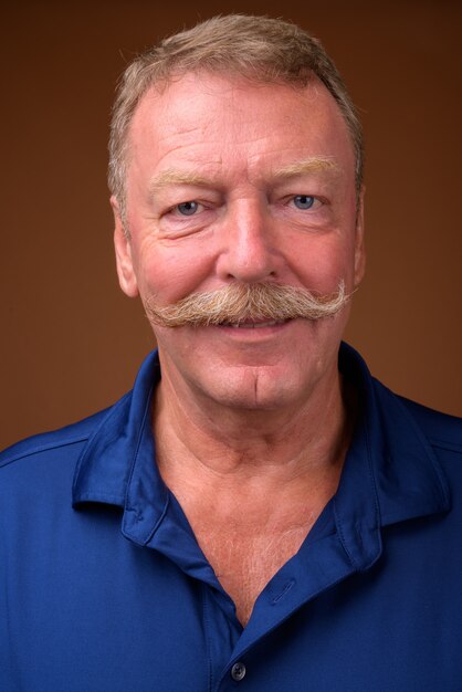 Foto de estúdio de um homem bonito sênior com bigode e camisa pólo azul contra marrom