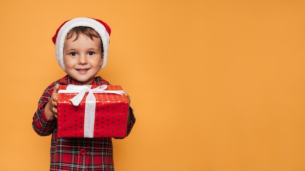 Foto de estúdio de um menino de pijama de Natal e um chapéu em um fundo amarelo com uma caixa de presente vermelha brilhante nas mãos. Um lugar para seu texto, publicidade.