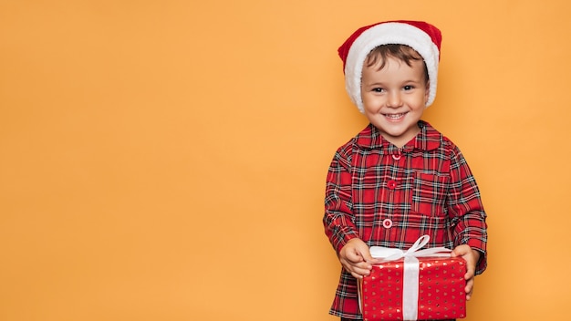 Foto de estúdio de um menino de pijama de Natal e um chapéu em um fundo amarelo com uma caixa de presente vermelha brilhante nas mãos. Um lugar para seu texto, publicidade.
