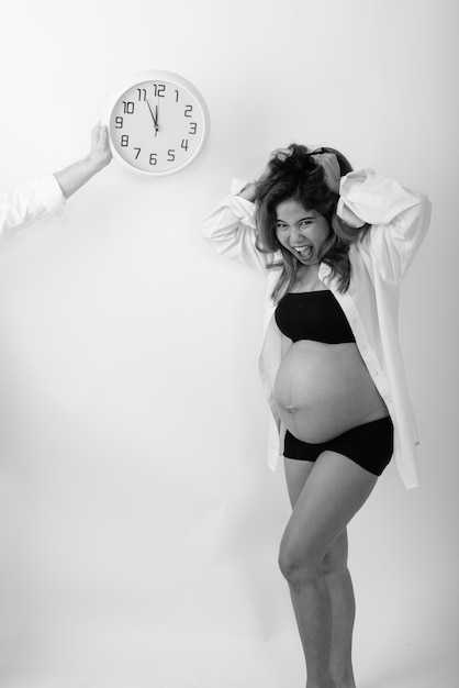 Foto de estúdio de uma jovem asiática grávida parecendo estressada com a contagem regressiva no relógio de parede contra a parede branca