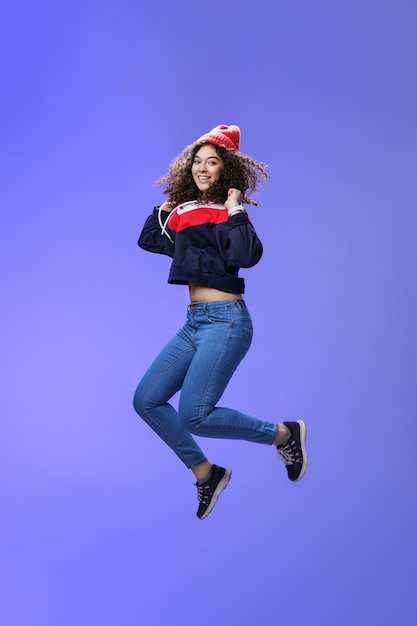 Foto de estúdio de uma linda garota com cabelo encaracolado em um gorro de moletom e tênis, pulando brincalhão e com cuidado ...