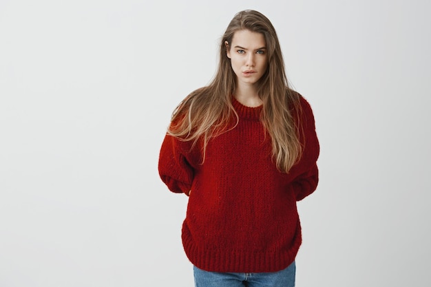 Foto de estúdio de uma mulher europeia atraente e preocupada, usando um suéter vermelho solto, mordendo o lábio e franzindo a testa