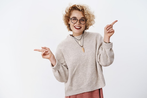 Foto de estúdio de uma mulher gay atraente e elegante, com corte de cabelo loiro encaracolado, sorrindo de forma positiva e despreocupada, apontando para a esquerda e para a direita, de lado, mostrando várias oportunidades e escolhas.