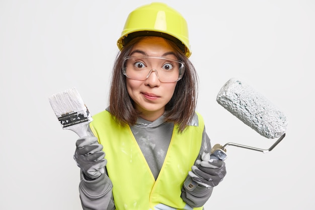 Foto de estúdio de uma mulher maravilhada, trabalhador industrial vestido de uniforme segurando pincel e rolo usa óculos de proteção, luvas de capacete envolvidas em trabalhos de construção, inspeciona a área