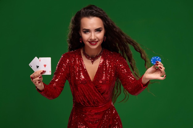 Foto de estúdio de uma mulher morena atraente em um vestido vermelho brilhante, com um penteado perfeito e maquiagem de noite. Ela mostra algumas cartas de baralho para a câmera, sobre um fundo verde. Cassino