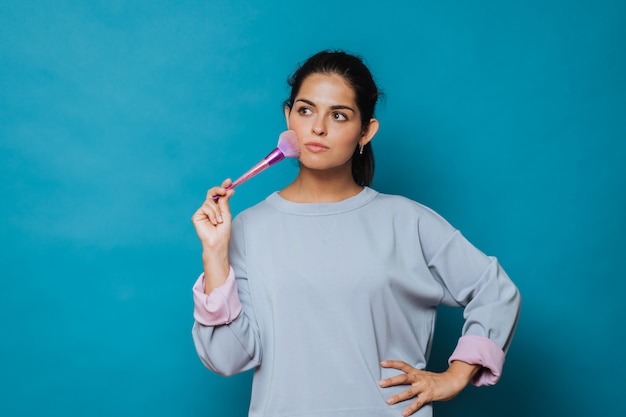 Foto de estúdio de uma mulher vestindo casual com um pincel para maquiagem em pó. Menina tocando sua bochecha com pincel, olhando de lado pensativamente.