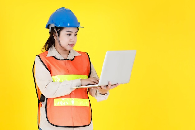 Foto de estúdio do gerente de contramestre de engenharia profissional feminino asiático usar capacete rígido e colete de segurança reflexivo ficar sorrindo segurando o computador portátil trabalhando em uma área industrial em fundo amarelo.