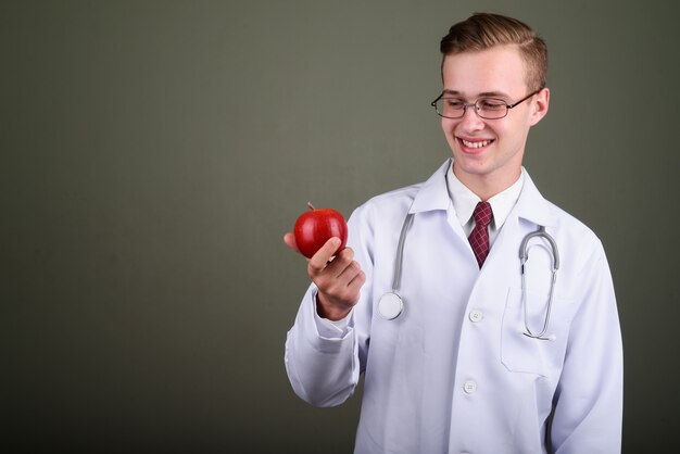 Foto de estúdio do médico jovem bonito usando óculos, segurando uma maçã vermelha contra um fundo colorido