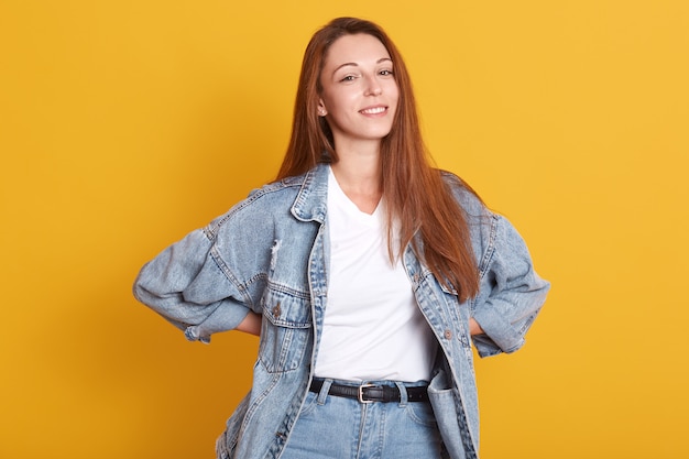 Foto de estúdio interior da moda jovem esbelta, mantendo as mãos na cintura, sorrindo sinceramente, vestindo jaqueta jeans, camiseta, jeans, com longos cabelos castanhos.