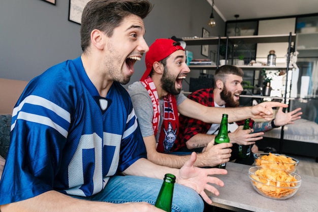 Foto de fãs jovens animados apontando o dedo e bebendo cerveja enquanto assistem a uma partida de esportes na sala de estar