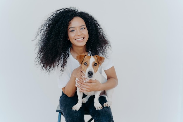 Foto de fêmea de pele escura feliz sendo para sempre junto com filhote de pedigree desfruta de lazer em casa senta-se na cadeira isolada sobre fundo branco Conexão humana e animal Confiança e amizade