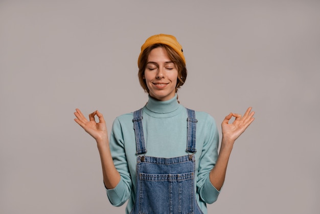 Foto foto de fofo hipster feminina meditação concentrada e relaxada. mulher branca usa macacão jeans e fundo de cor cinza com chapéu isolado.