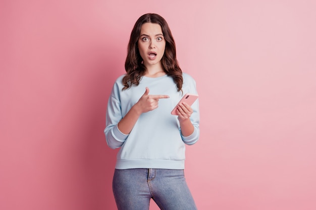 Foto de garota deslumbrante chocada com a boca aberta segurando o dispositivo de promoção de dedo direto do celular em fundo rosa