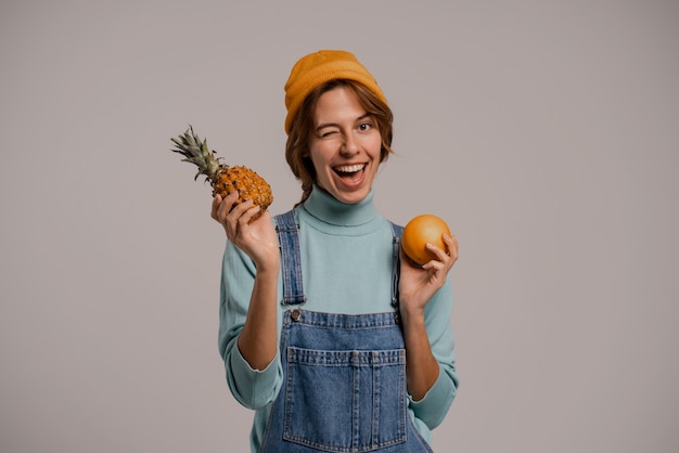 Foto foto de hipster feminino bonito brinca com abacaxi e laranja, parece saudável. mulher branca usa macacão jeans e fundo de cor cinza com chapéu isolado.