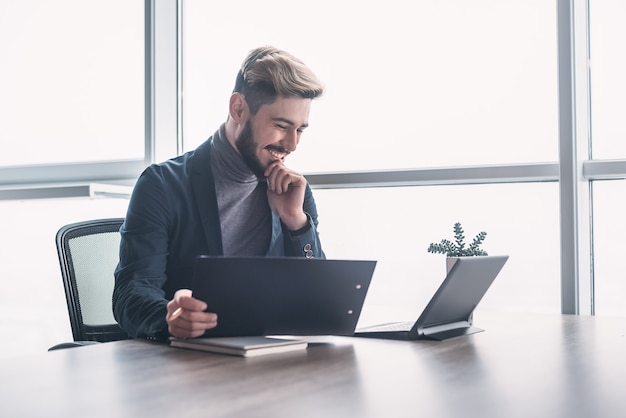 Foto de homem alegre com penteado moderno, vestido com jaqueta escura e gola olímpica cinza, usando o computador laptop enquanto compara os resultados com diagramas. coworking. olhe na tela.